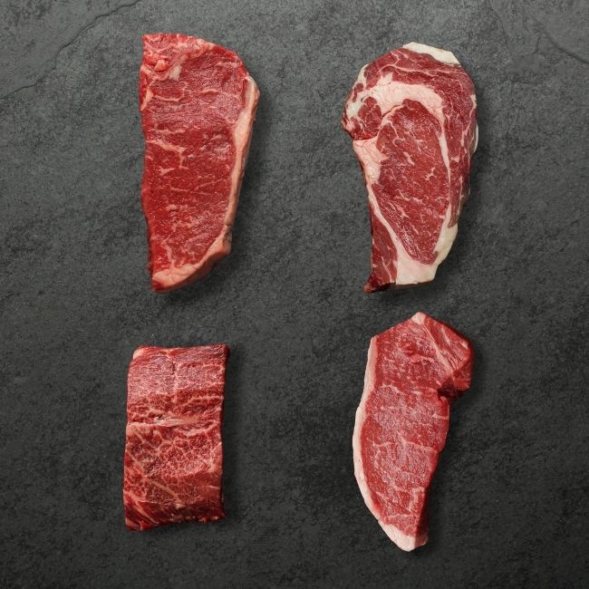 Overhead view of four different raw steaks, 1 flat iron steak, 1 rib eye steak, & 2 New York Steaks