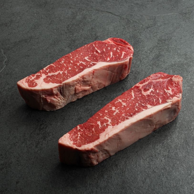 angled view of two raw, 16 oz new york strip steaks