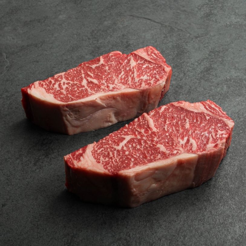 angled view of two richly marbled raw 16 oz new york strip steaks