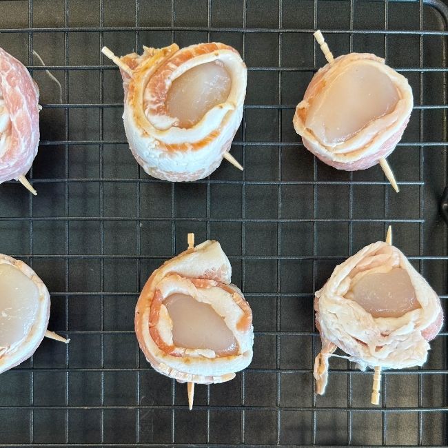 overhead view raw toothpick-skewed bacon-wrapped scallops on a wire rack-lined nonstick baking sheet