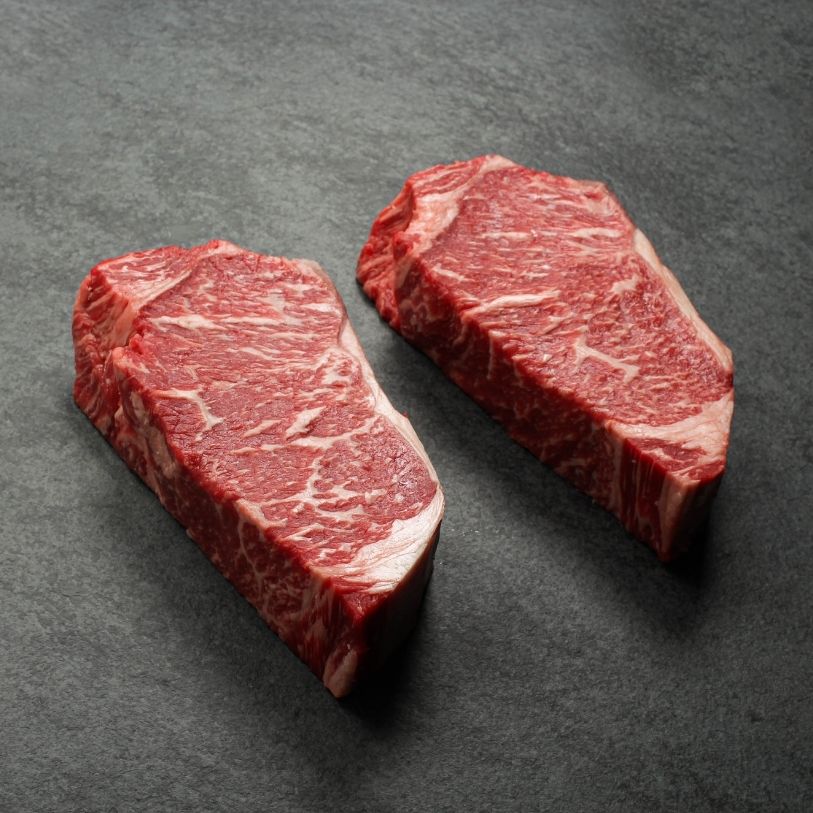angled view of two richly marbled raw 16 oz new york strip steaks