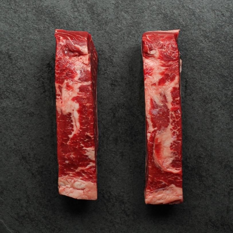 side view of two raw, 16 oz new york strip steaks