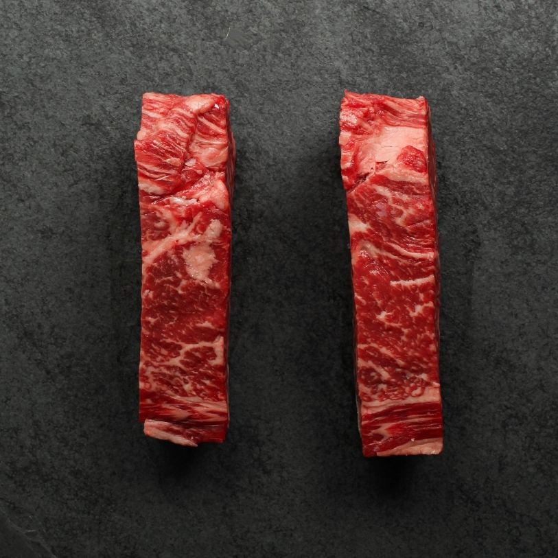 side view of two richly marbled raw 16 oz new york strip steaks