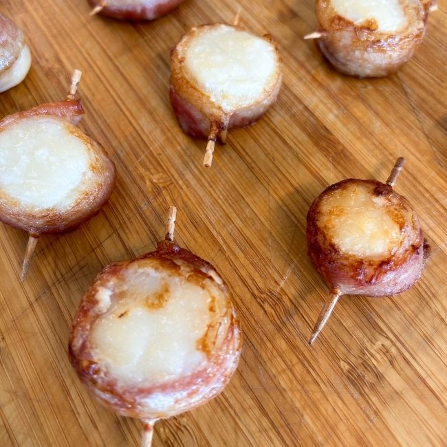 Cooked toothpick-skewered bacon-wrapped scallops on a wooden board