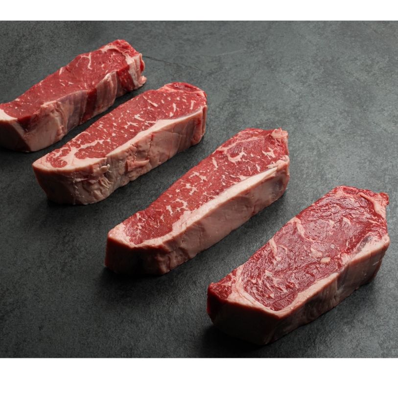 angled view of four raw, 16 oz new york strip steaks