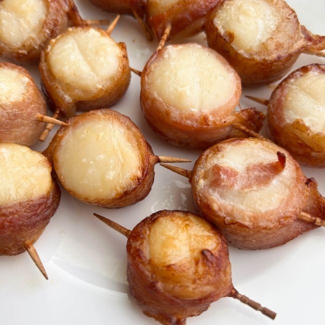 Cooked toothpick-skewered bacon-wrapped scallops on a white plate