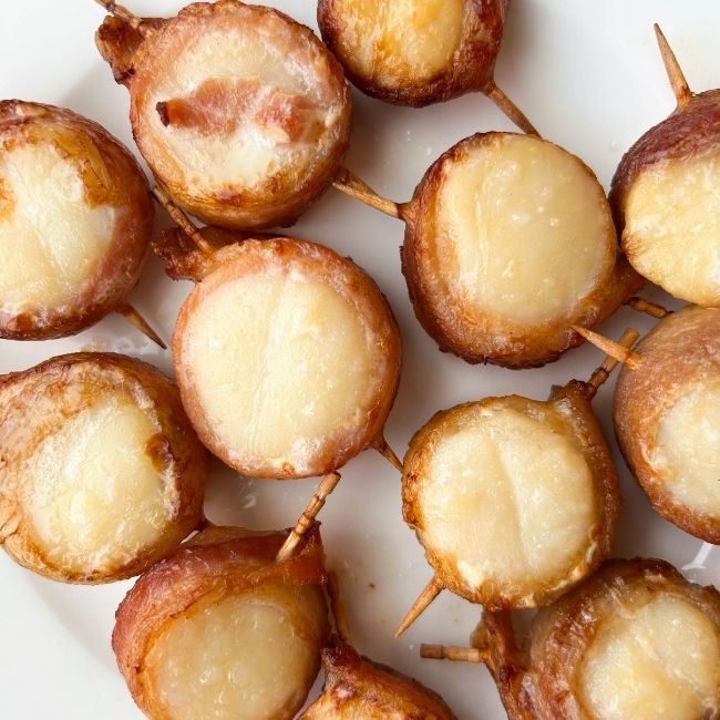 Cooked toothpick-skewered bacon-wrapped scallops on a white plate