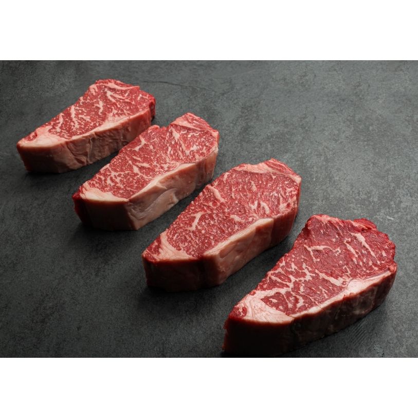 angled view of four richly marbled raw 16 oz new york strip steaks